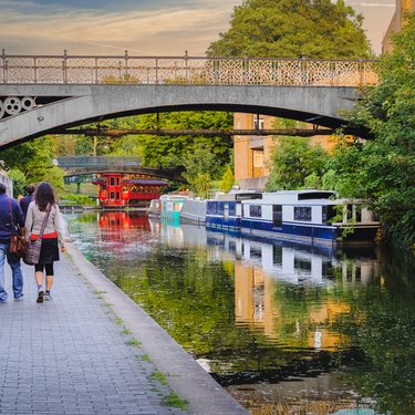 People walk on canal towpath under bridge with boats.